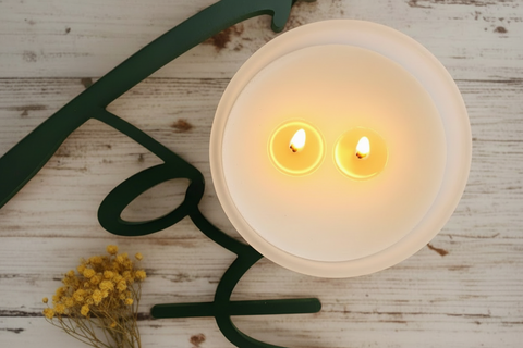 Candle in a white holder shaped like a face on a wooden surface with greenery.