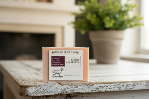 A bar of goat's milk bar soap in the Alaska Cranberry scent, displayed on a wooden shelf. The soap bar is a soft peach-pink color with a white paper label that features a black goat illustration and text detailing its organic and cruelty-free production in Alaska.