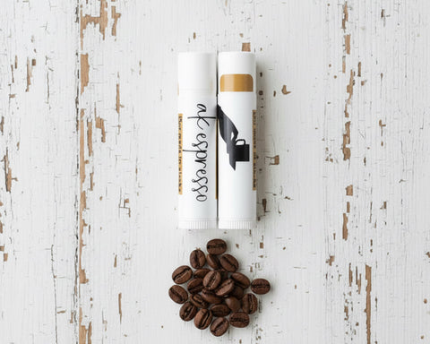Two white lip balm tubes labeled 'aK espresso' are arranged on a bed of roasted coffee beans on a distressed white wooden surface.