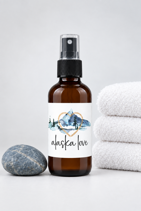 An amber glass spray bottle with a black pump and a white label reading 'alaska love' with a blue mountain illustration, standing on a light gray surface with folded white towels beside it.