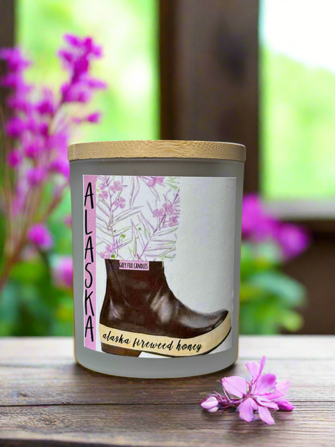 A frosted glass jar candle with a wooden lid, decorated with a graphic of a boot and fireweed, alongside a pink fireweed.