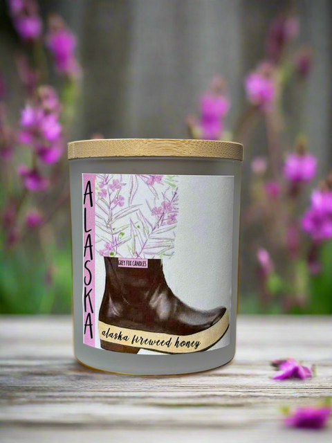 A frosted glass jar candle with a wooden lid, decorated with a graphic of a boot and fireweed, alongside a pink fireweed.