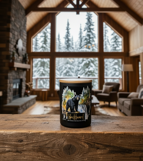 A black jar candle with a light wood lid sits on a rustic wooden table in a cozy, high-ceilinged log cabin living room. The candle features a graphic illustration of a black bear surrounded by snow ladened pine trees, with the text 'BlackBearD' printed below.