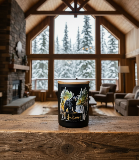 A black jar candle with a light wood lid sits on a rustic wooden table in a cozy, high-ceilinged log cabin living room. The candle features a graphic illustration of a black bear surrounded by snow ladened pine trees, with the text 'Black BearD' printed below.