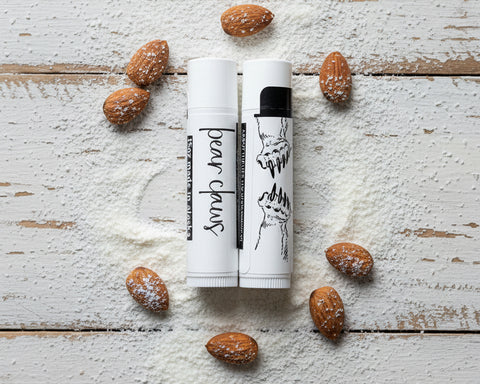 Two white lip balm tubes labeled 'bear claws' are arranged on a white wooden surface dusted with pastry powder, surrounded by whole almonds.