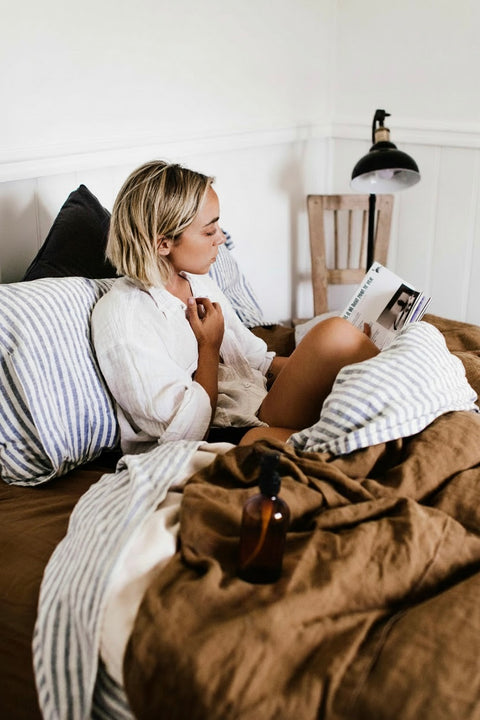 A woman with blonde hair sits on a bed reading a magazine, surrounded by brown and striped bedding in a bright bedroom. An amber bottle of linen spray is on the bed.