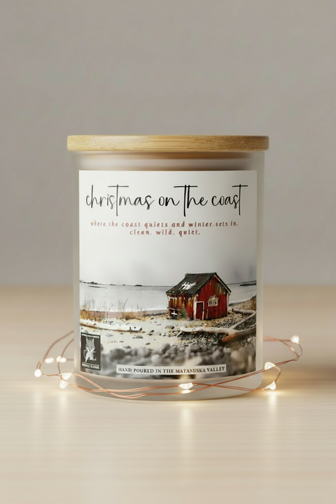 'Christmas on the Coast' featuring a small red cabin on a coastal beach with the alaska coastline in the background. The title is written in black script at the top, 'christmas on the coast' with red subtitle text in the middle and a 'Made in Alaska' logo at the bottom left.