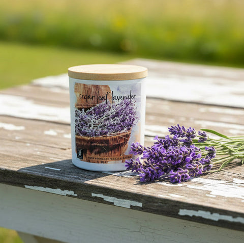 A white candle jar with a light wood lid sits on a weathered wooden table outdoors, next to a bouquet of purple lavender flowers. The candle label reads 'cedar leaf lavender'.