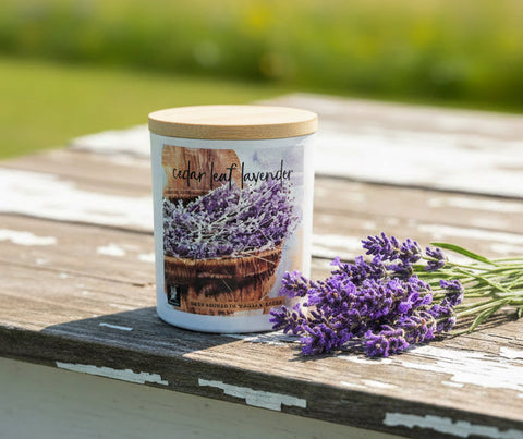 A white jar candle labeled 'cedar leaf lavender' with a light wood lid, displayed on a wooden shelf in front of other scented candles.