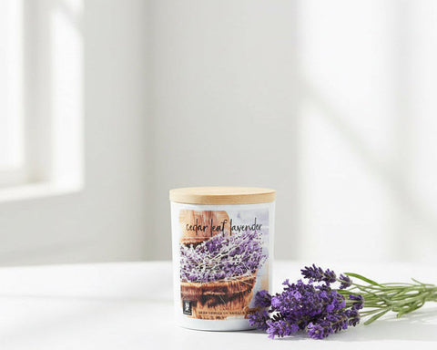 A white jar candle labeled 'cedar leaf lavender' with a light wood lid, displayed on a wooden shelf in front of other scented candles.