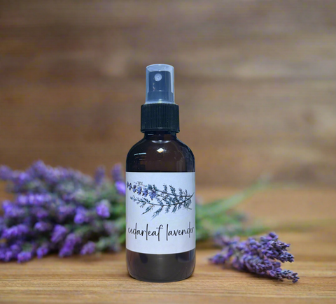 A dark amber glass spray bottle labeled 'cedar leaf lavender' with a white label and black spray cap, standing on a wooden surface with lavender sprigs in the background.