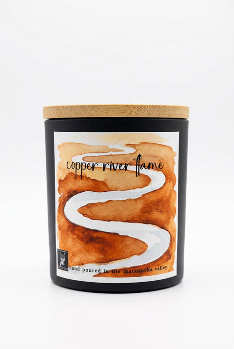 A matte black jar candle labeled 'copper river flame' with a watercolor-style copper and white winding river design on the front, by grey fox candles.