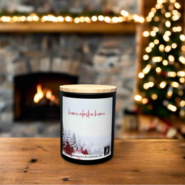 Candle with 'home alaska home' label in front of a fireplace and Christmas tree.