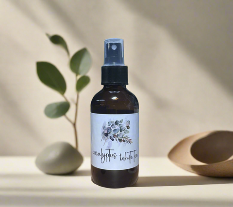 "Eucalyptus White Tea linen spray in amber bottle on a wooden table surrounded by lush green plants in a bright greenhouse, Grey Fox Candles"