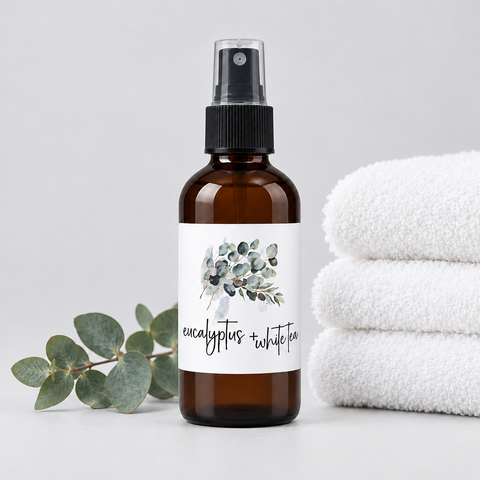 An amber glass spray bottle with a black spray top and a white label reading 'eucalyptus + white tea' with a watercolor eucalyptus leaf design, styled on a light gray surface with a sprig of eucalyptus leaves on the left and folded white towels on the right.