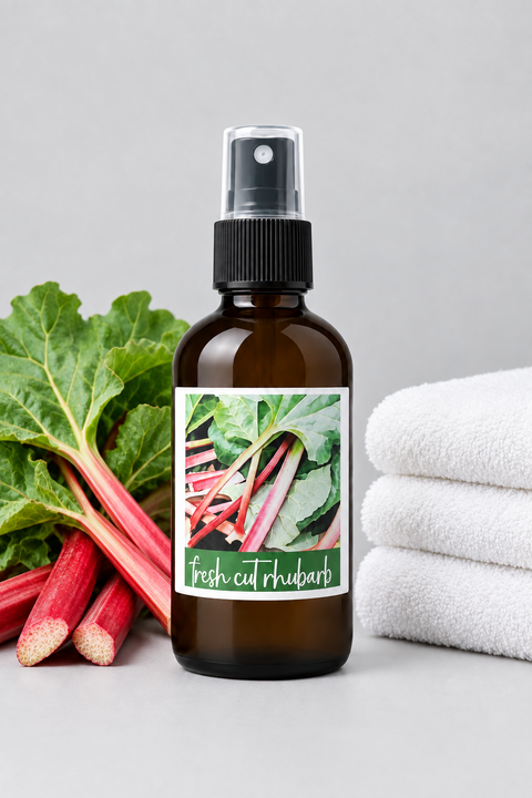 An amber glass spray bottle labeled 'Fresh Cut Rhubarb' stands on a light gray surface with fresh rhubarb stalks on the left and folded white towels on the right.