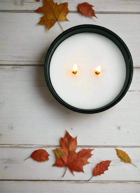 Top-down view of a lit Copper River Flame (maple bourbon) coconut wax candle with two cotton wicks burning in a matte black vessel, surrounded by fall maple leaves on a whitewashed wood surface, Grey Fox Candles