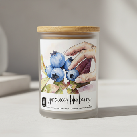 Watercolor-style candle label, showing a hand reaching for blueberries among leaves, with the text 'girdwood blueberry' and a small 'Made in Alaska' logo on a white background.