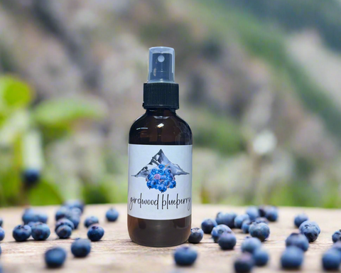 A dark amber glass spray bottle labeled 'girdwood blueberry' with a white label and blueberry illustration, standing on a wooden surface surrounded by scattered blueberries with a blurred outdoor background.