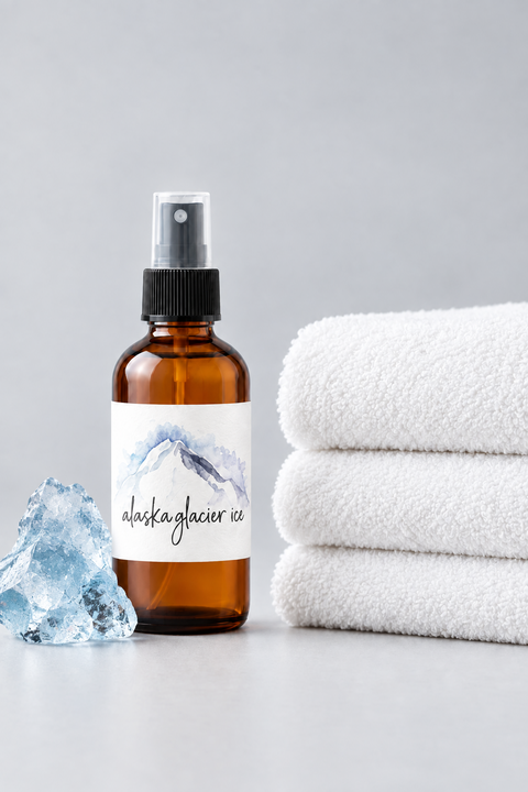 An amber spray bottle labeled 'alaska glacier ice' stands on a light gray surface next to a stack of three folded white towels and a blue crystal, against a soft light gray background.