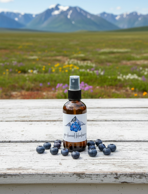 A dark amber glass spray bottle labeled 'girdwood blueberry' with a white label and blueberry illustration, standing on a wooden surface surrounded by scattered blueberries with a blurred mountain background.