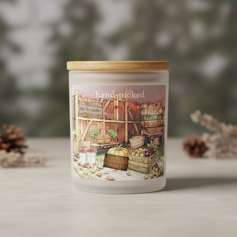 Vintage-style illustrated sign and rustic market scene with the words 'hand-picked' at the top. The artwork shows wooden barn fencing, produce crates, buckets of berries, a pumpkin, and signs including 'grey fox candles, and 'FRESH ALASKAN PRODUCE'