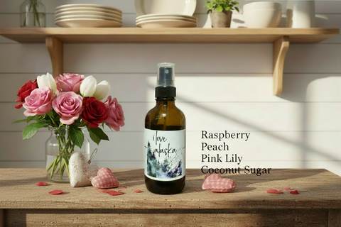 Bottle of 'I Love Alaska' linen spray on a wooden surface with flowers and shelves in the background