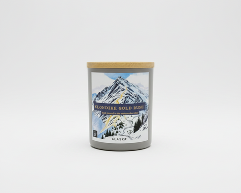A frosted white glass jar candle with a light bamboo lid and a detailed label depicting a snowy mountain scene. The label reads 'KLONDIKE GOLD RUSH' and 'ALASKA'.