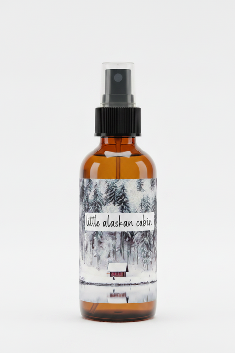 An amber glass spray bottle with a black pump dispenser and a winter-themed label reading 'little alaskan cabin', shown on a plain white background.