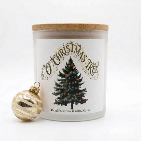 A frosted glass jar candle with a bamboo lid and a white label reading 'O Christmas Tree' with a decorated tree illustration. A gold-and-white striped ornament sits beside the candle on a clean white background.