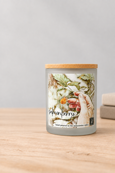 Candle with salmonberry watercolor decorative label on a wooden surface in front of a stone fireplace, by grey fox candles