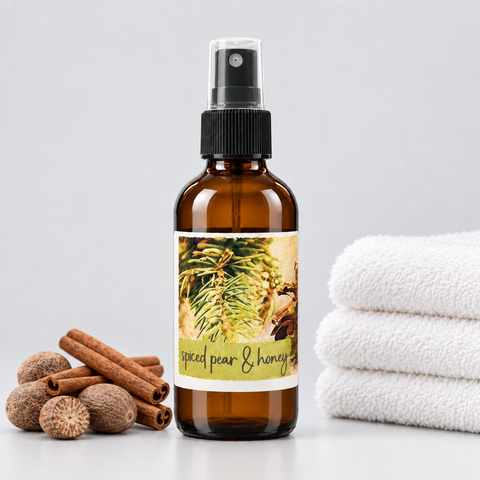 An amber glass spray bottle with a black pump and a decorative label reading 'spiced pear & honey,' styled on a light gray surface with cinnamon sticks and nutmegs on the left and folded white towels on the right.