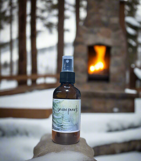Snow Pearls linen and room mist in amber spray bottle with snowy spruce tree label, held up outdoors in front of a snow-covered stone fireplace, Grey Fox Candles"