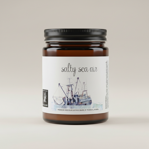 An amber glass jar candle with a black metal lid and a white paper label reading 'salty sea air' above a watercolor illustration of a fishing boat. The label also includes a small black square logo and text indicating premium scented beeswax lotion made in Wasilla, AK