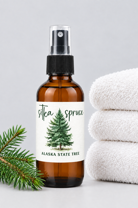 An amber glass spray bottle labeled 'sika spruce ALASKA STATE TREE' with a pine tree illustration, styled on a light gray surface with a green pine branch and a stack of white towels.