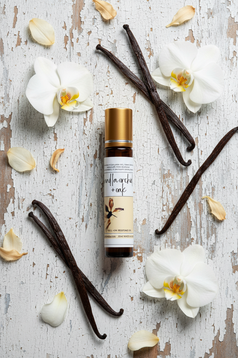 A rectangular product label for greyfoxcandles vanilla orchid + oak roll-on perfume oil. The label has a cream background with black text and a central illustration of a vanilla orchid flower. The label includes product details, ingredients, and the brand made in alaska by grey fox candles.