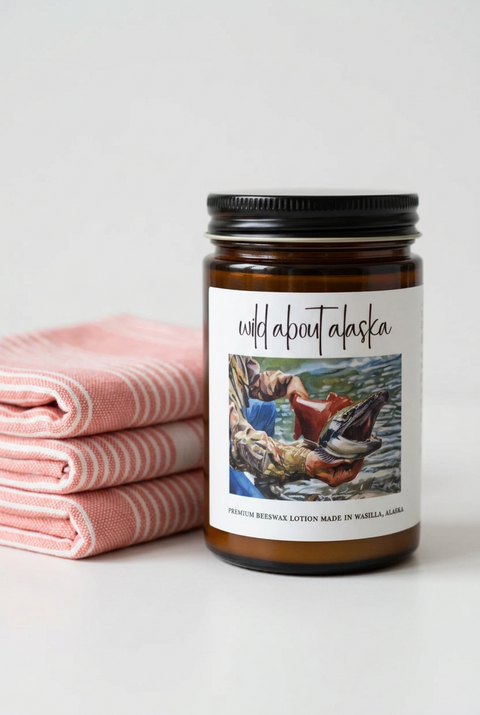 An amber glass jar of Wild About Alaska premium beeswax lotion with a black metal lid and a white label featuring a fishing illustration, set against a neutral gray background. To the right are salmon + white colored pinstripe turkish towels.