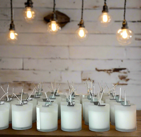 A group of frosted glass candle jars filled with white wax, arranged on a wooden surface against a rustic white-painted wood wall with hanging Edison bulbs. grey fox candles sells wholesale