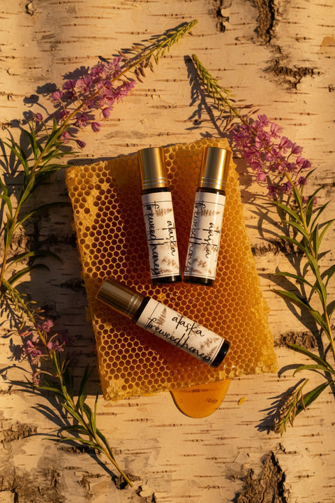 Three small perfume roller bottles with gold caps and white labels are arranged on a honeycomb honey block over a birch tree background, surrounded by pinkish purple fireweed flowers and green leaves. fragrance is alaska fireweed honey, made in alaska by grey fox candles.