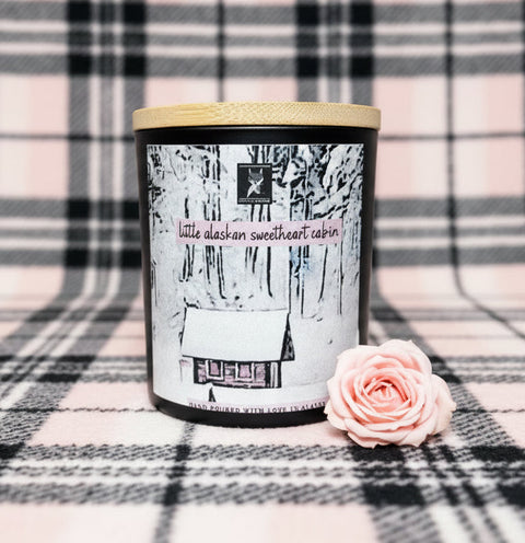 Candle with a label featuring a cabin design on a plaid background with a pink rose.