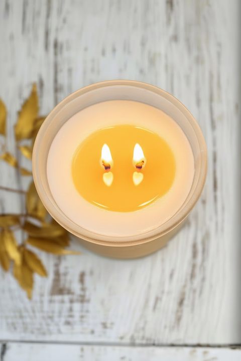 A lit cream-colored candle in a frosted glass jar with a beige rim, placed on a distressed white wooden surface with dried gold leaves in the corner.