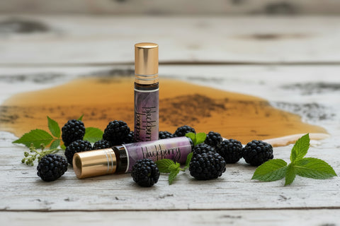 two small perfume roller bottles with gold caps and beautiful labels are arranged on a birch tree background, surrounded by fresh ripe blackberries and a spilled bourbon; green mint sprigs are scattered casually around the bottle. made in alaska by grey fox candles.