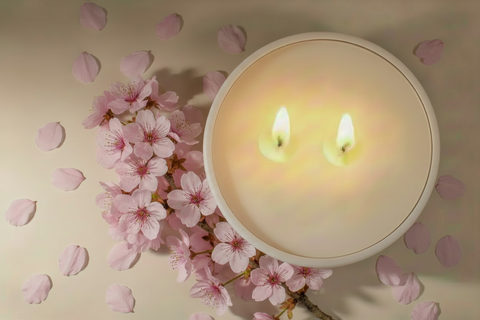 A round white candle with two lit wicks, styled on a beige surface with scattered pink cherry blossom petals and a cherry blossom branch.