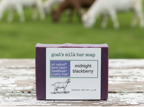 A bar of goat's milk soap labeled 'midnight blackberry' sits on a weathered white wooden table, with a blurred green field and grazing goats in the background.