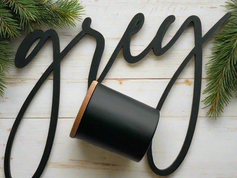 A black matte glass jar candle with a wooden wick, placed on a white surface with greenery in the background, and a stylized black text saying 'grey'.
