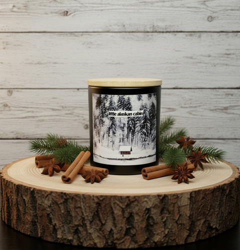 Candle labeled 'Little Alaskan Cabin' with cinnamon sticks and pine branches on a wooden surface.