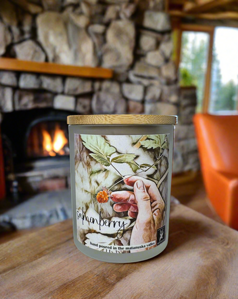 Candle with salmonberry watercolor decorative label on a wooden surface in front of a stone fireplace.