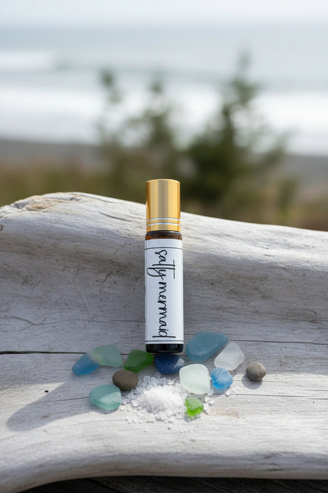 A small amber glass roller bottle with a gold cap and a white label reading 'salty mermaid' fragrance stands on a weathered driftwood log outdoors, surrounded by sea glass stones and a small pile of white salt. made in alaska by grey fox candles