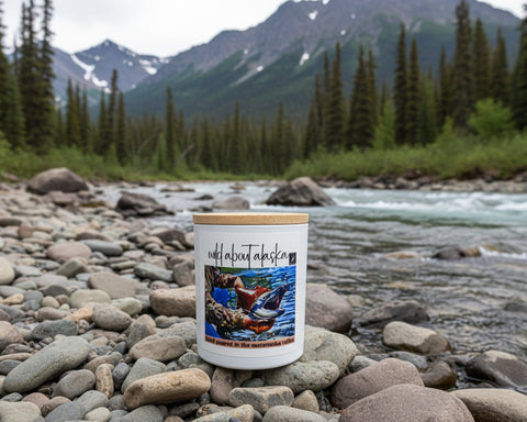 candle with a nature-themed label on pebbles near a river and mountains