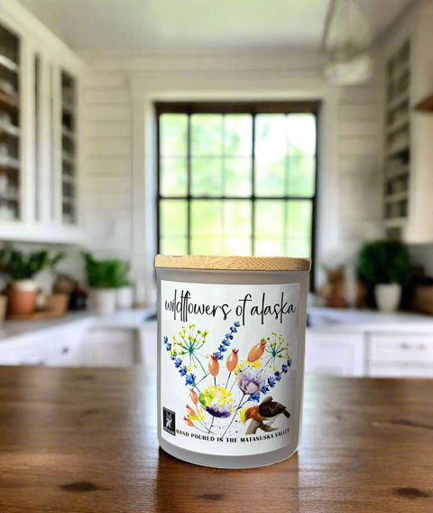 Candle labeled 'Wildflowers of Alaska' on a wooden table in a kitchen.